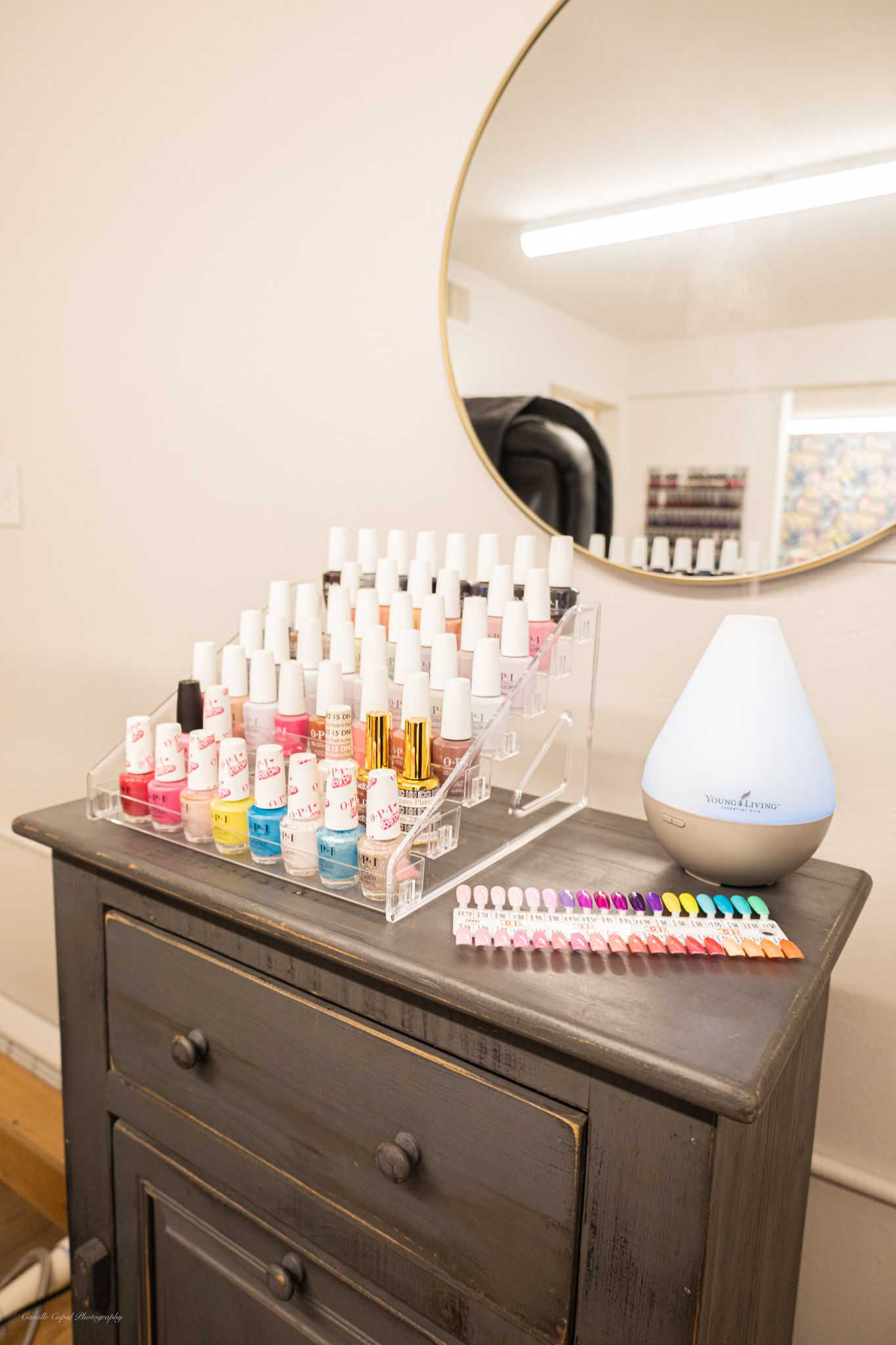 Display of colorful nail polishes and an aroma diffuser on a dark wooden dresser with a round mirror.